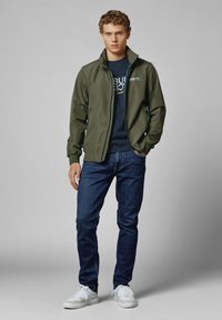 Alyvuogių žalia lengva striukė su stovinčiu apykakle, užtrauktukais, derinama su tamsiai mėlynomis marškinėliais ir tamsiai mėlynomis džinsais, papildyta baltais sportiniais bateliais.