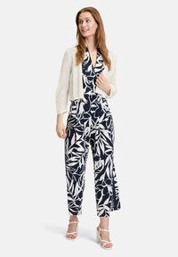 Marineblauwe jumpsuit met wit loofpatroon, wijde pijpen en een V-hals. Gecombineerd met een beige kort geknipt cardigan en witte block-heeled sandalen.