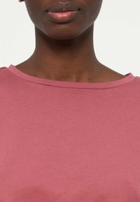 Κοντινή λήψη ενός ατόμου που φοράει ροζ μπλουζάκι με στρογγυλή λαιμόκοψη, εμφανίζοντας τα χείλη, τον λαιμό, την κλείδα και έναν μικρό σπίλο στη δεξιά πλευρά.