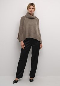 Bež kudumi ponšo, millel on kõrge krae, külgadel nupud ja lahtine lõige, koos mustade laiade pükstega ja mustade kontsakingadega.