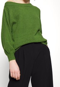 Groene gebreide trui met een off-shoulder ontwerp, lange mouwen en geribde manchetten, gecombineerd met zwarte hoge taillebroeken.