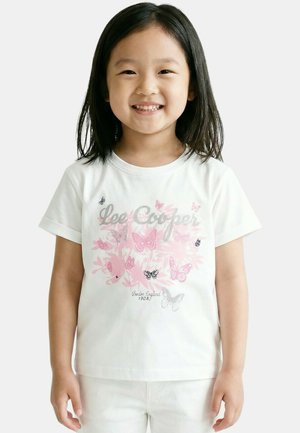 Jeune fille souriante aux cheveux noirs et raides portant un t-shirt blanc avec des papillons roses et gris ainsi que le texte "Lee Cooper" et un pantalon blanc.