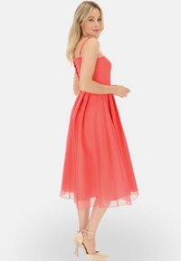 Koronkowa sukienka w kształcie litery A w kolorze koralowym z cienkimi ramiączkami, dopasowanym gorsetem i zwiewnym, przezroczystym dołem. W komplecie z cielistymi butami i delikatną bransoletką.