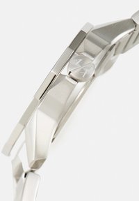 Ura iz nerjavečega jekla z geometrijsko zasnovo ohišja, poliširano površino in stranskim gumbom z vgraviranima črkama "A" in "X".