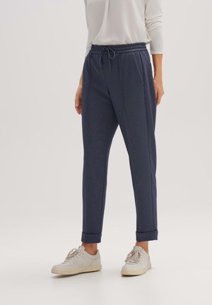 Marineblauwe tapse broek met een elastische tailleband en een trekkoord. Kenmerken zijn frontplooien en omgeslagen manchetten. G gedragen met witte sneakers.