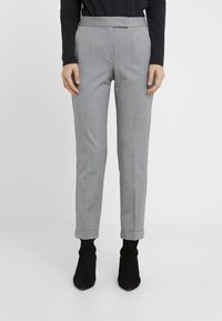 Persoon in grijze gepaste enkellange broek met omslagen, een zwart langarmtop en zwarte suède enkellaarzen tegen een witte achtergrond.