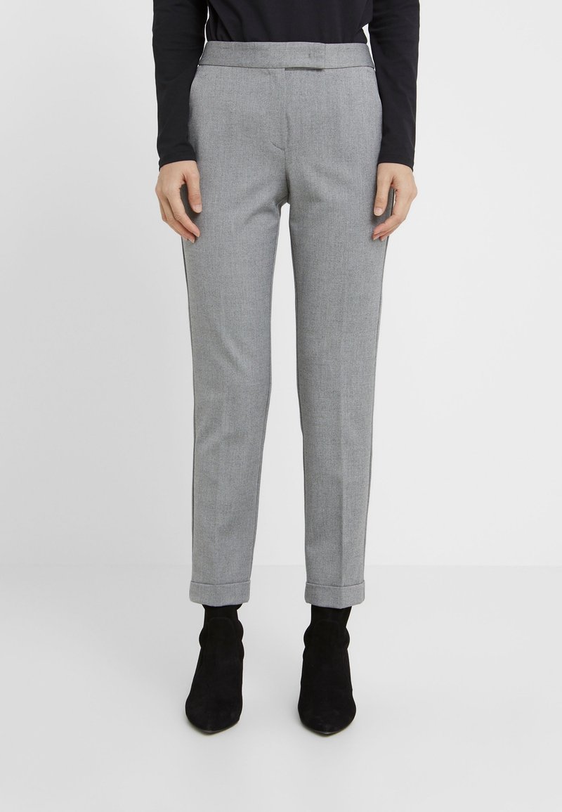Persoon in grijze gepaste enkellange broek met omslagen, een zwart langarmtop en zwarte suède enkellaarzen tegen een witte achtergrond.