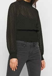Blusa nera trasparente con pois testurizzati, colletto alto plissettato e polsini elasticizzati. Abbinata a jeans scuri, che evidenziano uno stile aderente.