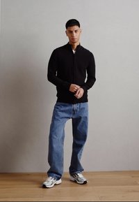 Svart zip-up genser, løse blå jeans og hvite sneakers. Modellen står på tregulv med grå vegg i bakgrunnen.