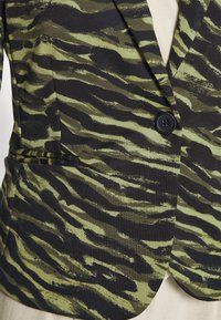 Blazer a rayas de tigre negro y verde que presenta un solo botón negro, tela texturizada y un bolsillo frontal en el lado izquierdo.