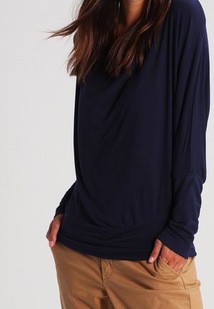 Frau trägt ein locker sitzendes, marineblaues Langarmshirt und hellbraune Hose, mit den Händen in den Taschen vor einfachem Hintergrund.