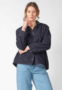 Kobieta o jasnobrązowych włosach, ubrana w czarną sztruksową kurtkę zapinaną na guziki oraz jasnoniebieskie dżinsy, pozująca z jedną ręką na podbródku, a drugą skrzyżowaną na piersi.