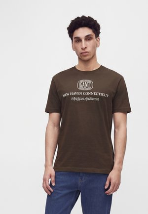 Jeune homme aux cheveux bouclés foncés portant un t-shirt marron GANT avec un texte blanc et un jean bleu, debout sur un fond blanc uni.
