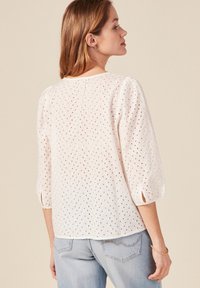 Weiße Bluse aus Lochstickerei mit Puffärmeln, einem runden Ausschnitt und kleinen Ausschnitten. Kombiniert mit hellblauen Jeans, die eine lässige Silhouette zeigen.