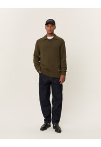 Olivgrön stickad tröja med krage och tre knappar, tillsammans med mörkblå jeans och svarta skor. Modellen står mot en neutral bakgrund.