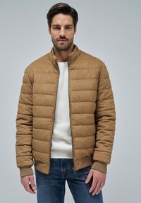 Chaqueta marrón acolchada con cuello alto, cierre de cremallera frontal, puños de canalé y textura suave, llevada sobre un suéter blanco y unos jeans azules.