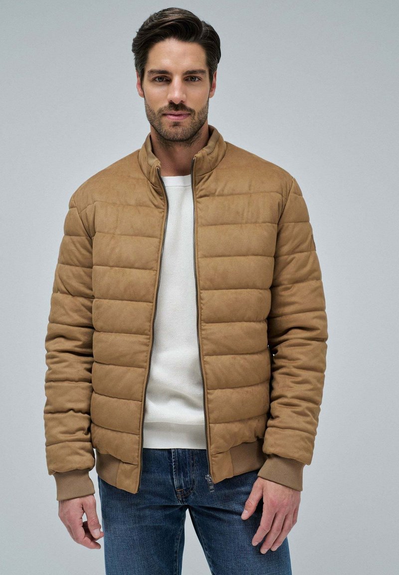 Chaqueta marrón acolchada con cuello alto, cierre de cremallera frontal, puños de canalé y textura suave, llevada sobre un suéter blanco y unos jeans azules.