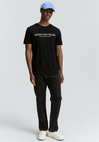 Musta puuvillapaita, jossa on "DENIM TOM TAILOR" -logo ja "ESTABLISHED 2007" harmaana, yhdistetty mustiin farkkuihin ja valkoisiin lenkkareihin, päällä vaaleansininen lippis.