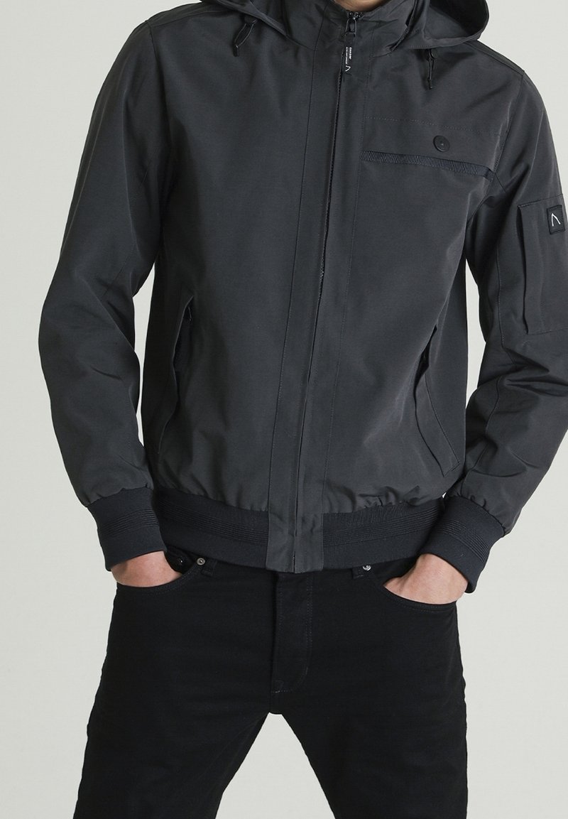 Personne portant une veste noire à capuche zippée avec poignets et taille élastiques, mains dans les poches, associée à un pantalon noir sur un fond uni.