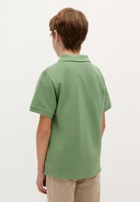 Polo shirt vert à finition texturée, à manches courtes et avec un col plat. Porté avec un pantalon beige clair, vu de dos.