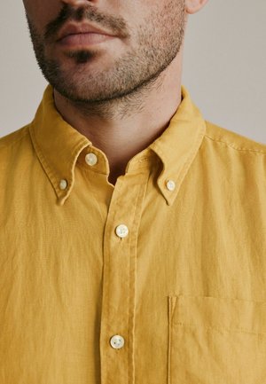 Close-up of a man’s lower face and neck wearing a mustard yellow buttoned shirt with a collar and chest pocket.