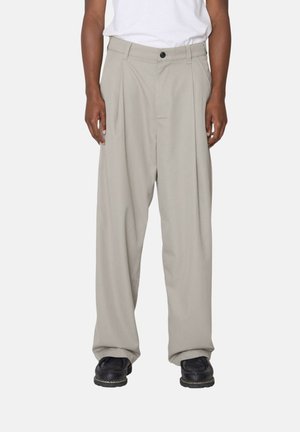 Mann trägt beige, weit geschnittene plissierte Hosen, weißes T-Shirt und schwarze Schuhe, steht vor einem einfarbigen Hintergrund.