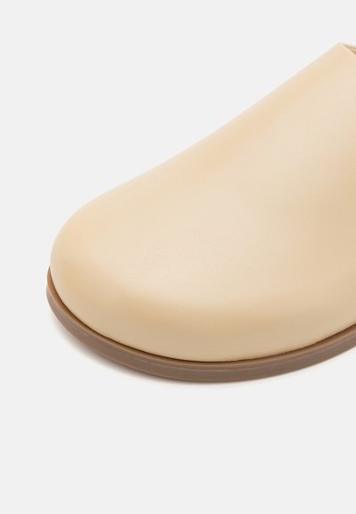 Botte beige à enfiler avec une texture lisse, un bout arrondi et une semelle en caoutchouc brun clair. Design minimaliste, sans motifs ni ornements visibles.