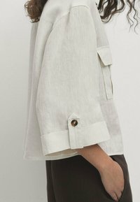 Light beige linen shirt with a relaxed fit, roll-up sleeves, large patch pocket, and button details on cuffs, showcasing a subtle texture.