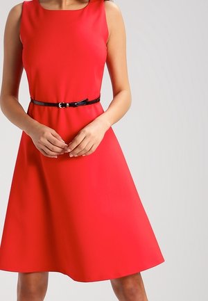 Robe rouge sans manches avec une fine ceinture noire à la taille, portée par une personne se tenant devant un fond clair.