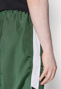 Rechterhand rustend naast groene korte broek met witte en zwarte zijkantstreep, zichtbaar deel van zwart shirt boven de taille.
