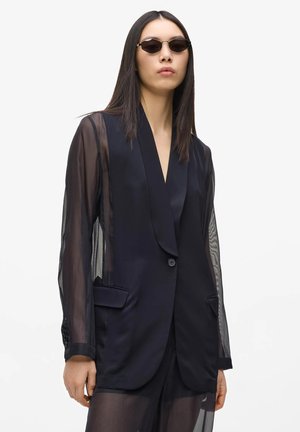 Kvinde iført sort transparent langærmet blazer med satin revers og sorte ovale solbriller, stående mod en ensfarvet hvid baggrund.