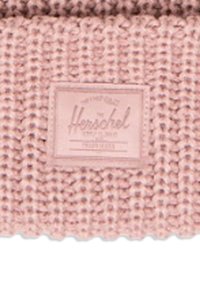 Tejido de punto rosa claro con un patrón texturizado; presenta una etiqueta rectangular en relieve con el logo de Herschel en el centro.