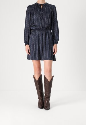 Frau trägt ein marineblaues langärmliges Kleid mit kleinen weißen Punkten, kombiniert mit knielangen dunkelbraunen Cowboystiefeln.