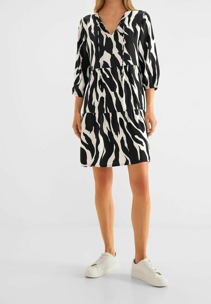 Žena v šatech s černobílým zebrovým vzorem a tříčtvrtečními rukávy, oblečená v bílých platformových teniskách, stojí na jednobarevném pozadí.