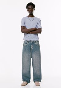 Un giovane uomo in piedi con le braccia incrociate, che indossa una maglietta grigia chiara, jeans larghi blu sbiaditi, sneakers beige e gioielli in argento.