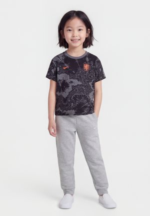 Barn står og smiler, iført en svart og gråmønstret Nike-T-skjorte med oransje logoer, grå joggebukser og hvite slip-on-sko.