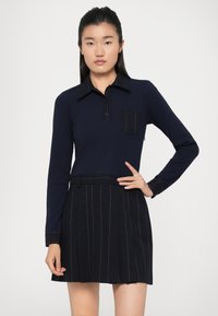 Marineblå langermet polo med svart krage og lomme, kombinert med et svart plissé skjørt med hvite striper.