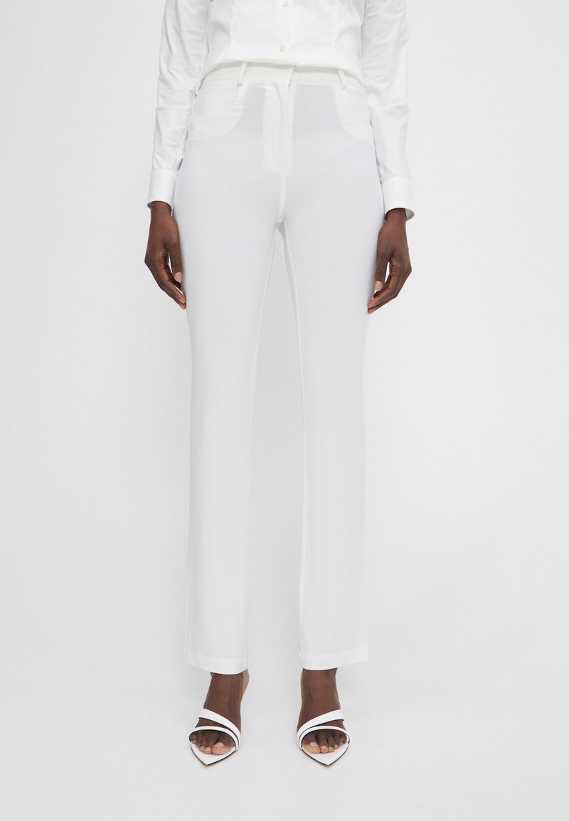 Pantalons tailleurs blancs en tissu lisse, avec une taille plate, des poches latérales et une coupe droite. Portés avec des talons ouverts blancs.
