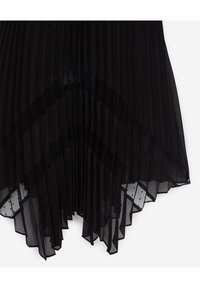 Falda negra plisada con superposición transparente, con un diseño geométrico y lunares a lo largo del dobladillo. Tela ligera con textura suave.