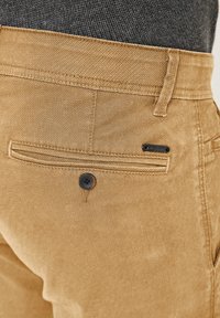 Beige cotton trousers with a textured fabric, featuring a back pocket with a black button and a subtle logo tag above the pocket.