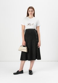 Camiseta blanca con "Karl" en lentejuelas coloridas, combinada con una falda negra brillante de longitud media con botones en un lado. Bolso beige.