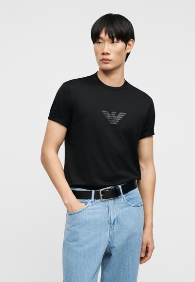 Νεαρός άνδρας με μαύρο μπλουζάκι με γκρίζο λογότυπο, ανοιχτόχρωμα μπλε τζιν, μαύρη ζώνη, στέκεται με το ένα χέρι στην τσέπη, κοιτώντας προς το πλάι.