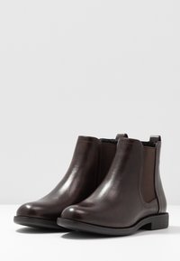 Botas Chelsea de cuero marrón con paneles laterales elásticos, puntera redondeada y tacón bajo. Textura suave con detalles mínimos.