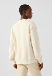Sudadera color crema con un corte relajado, mangas largas, puños y dobladillo acanalados, y textura suave, vista desde la parte posterior.