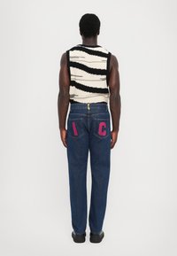 Jean en denim avec une coupe droite, de couleur bleu foncé, présentant des lettres brodées en violet "I" et "C" sur les poches arrière. Débardeur en tricot texturé.