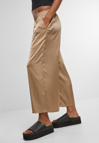 Light brown satin wide-leg pants with elastic waistband and side pockets, paired with black platform slide sandals. Smooth texture, loose fit.