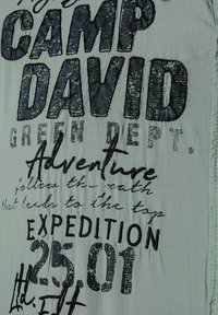 Groene stof met gedurfde zwarte bedrukte tekst, waaronder "CAMP DAVID" en "ADVENTURE." Bevat geprofileerde accenten en verschillende letterstijl.