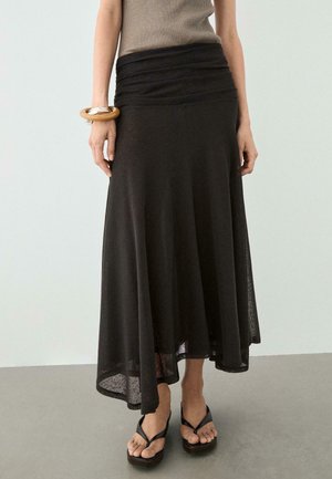 Frau trägt einen schwarzen Midi-Rock mit hoher Taille, beige ärmelloses Top, Holzarmreif und schwarze Plateau-Sandalen mit Zehenriemen.