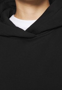 El sudadera con capucha negra cuenta con un tejido suave, dobladillo acanalado y una capucha envolvente. Se ve una camiseta blanca de cuello redondo debajo.