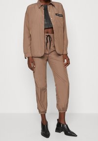 Žena oblečená v hnědém sako na knoflíky a odpovídajících jogger kalhotách, s černým zkráceným koženým topem a černými koženými kotníkovými botami na podpatku.